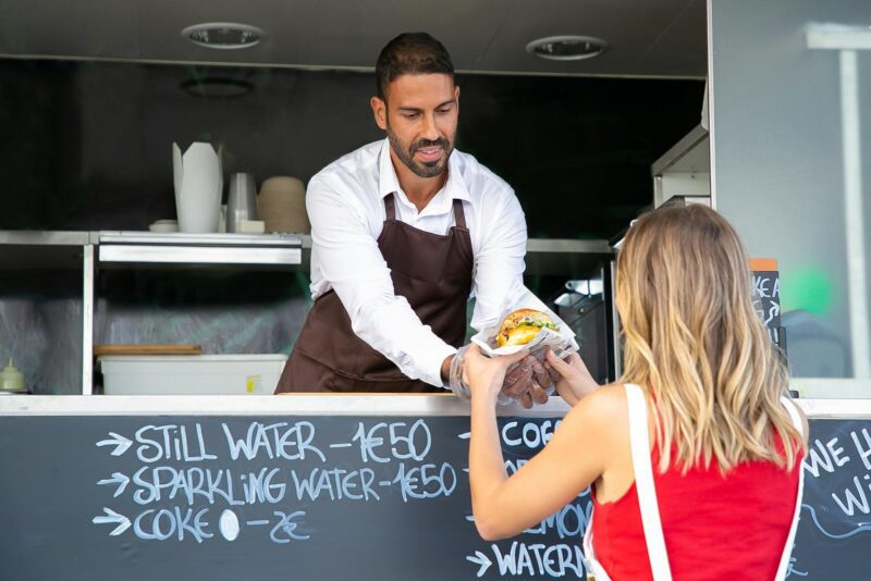 Positive ethnic cook in apron standing at counter in food truck and giving delicious hamburger to anonymous woman customer in daytime